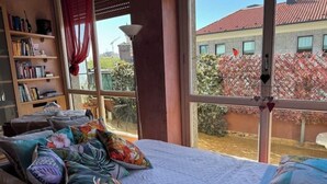 Ein Schlafzimmer mit großem Fenster, einer Bücherwand und einem Bett mit blumigen Kissen.