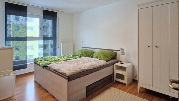 A modern bedroom with a bed, bedside table, wardrobe, and a large window. - Furnished apartment near Plauen's old market square with sunny balcony (Plauen)