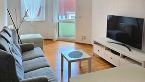 A modern living room with a grey sofa, a small table, a TV on a stand, and a large window with curtains. - Furnished apartment near Plauen's old market square with sunny balcony (Plauen)