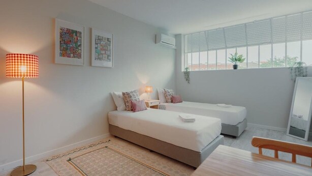 A bedroom with two beds, a standing lamp, a window with blinds, and a mirror. - Colorful Studio 1 Municipal Pool by Vacationy (Porto)