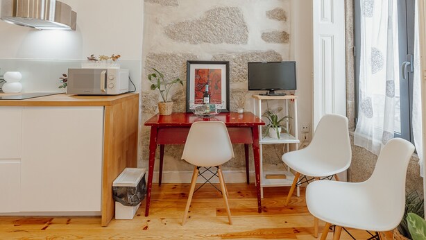A room with a wooden floor, a stone wall, a wooden table with chairs, a microwave, a framed picture, a television, and a window with curtains. - Marques 1243 Studio by Vacationy (Porto)