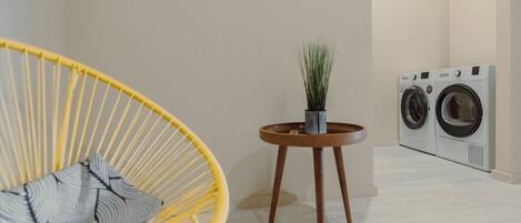 A room with a yellow chair, a small wooden stool with a plant, and a washing machine.