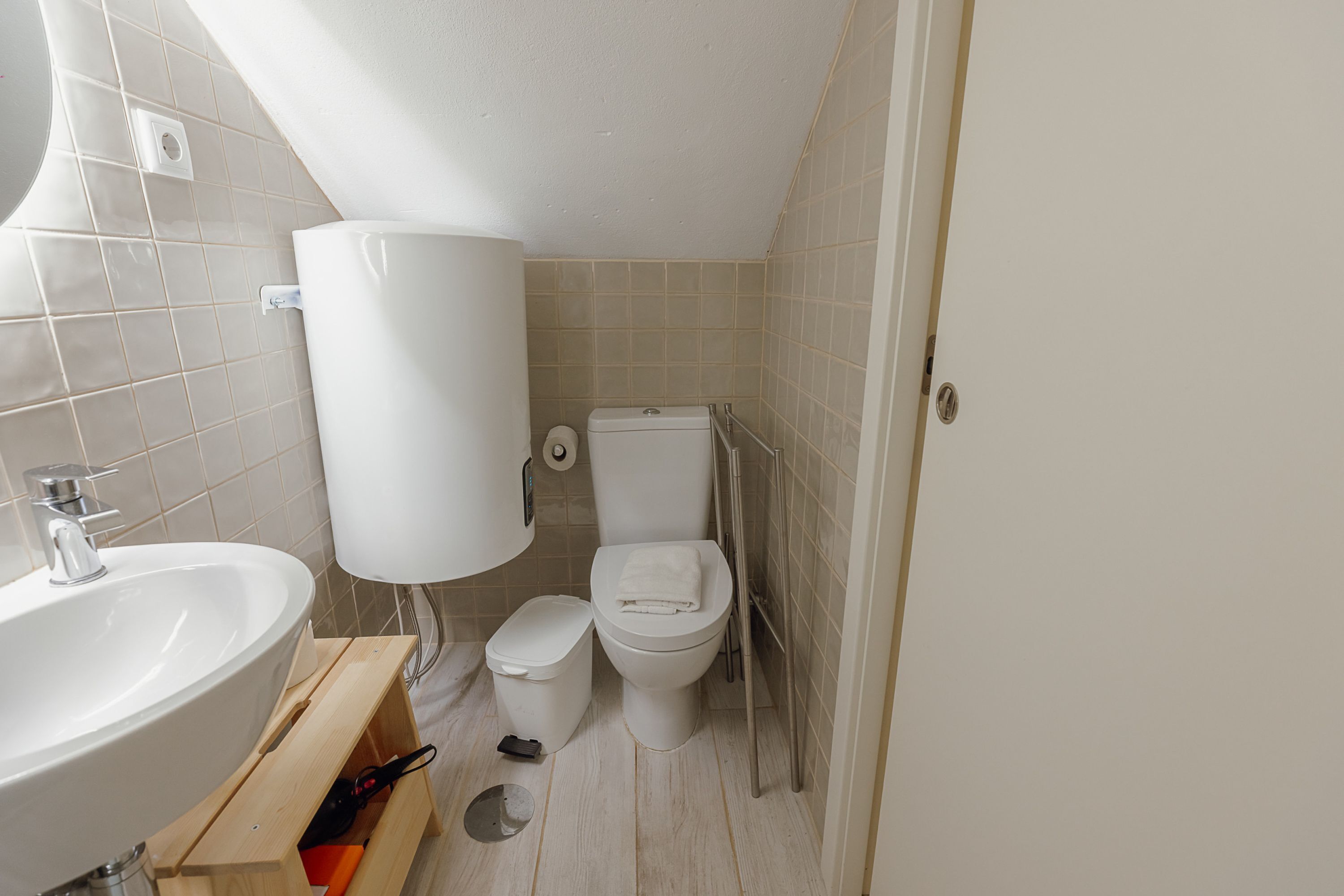 Egy kis fürdőszoba, amelyben mosdókagyló, WC és egy fehér WC-papírtartó található.