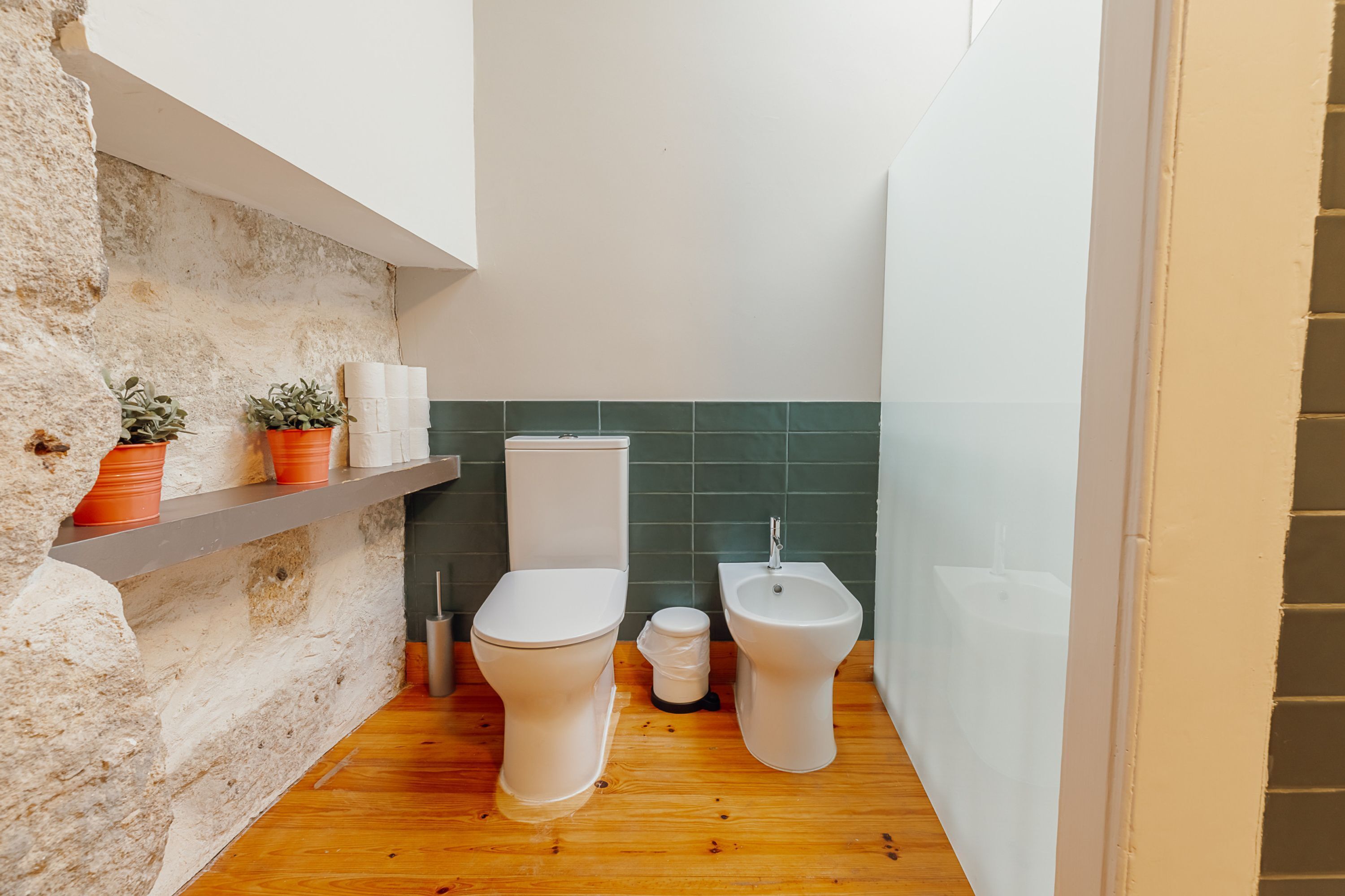 A compact bathroom with a white toilet, a bidet, and a small trash bin. There are two potted plants on a shelf and a green tiled backsplash.