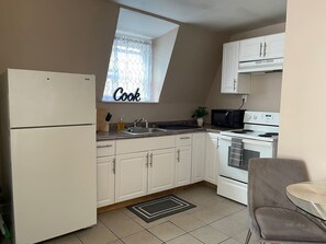 Eine kompakte Küche mit weißen Schränken, einem Kühlschrank, einer Mikrowelle, einem Backofen und einem Spülbecken. Über einem kleinen Fenster hängt ein Schild mit der Aufschrift 'Cook'.