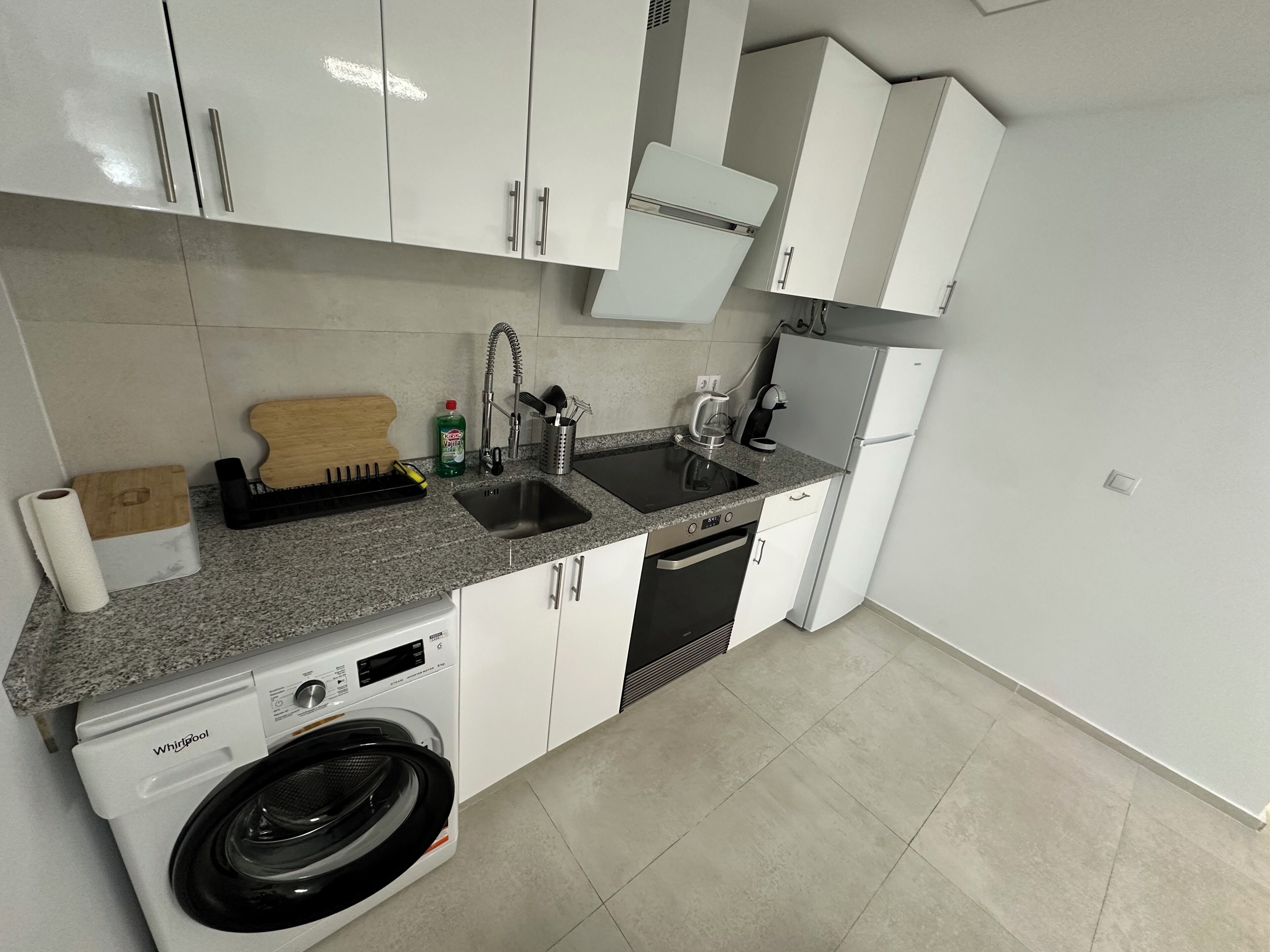Cocina compacta con armarios blancos, lavavajillas integrado, lavadora, pileta con grifo y cafetera.