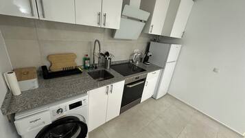 Cocina compacta con armarios blancos, lavavajillas integrado, lavadora, pileta con grifo y cafetera.