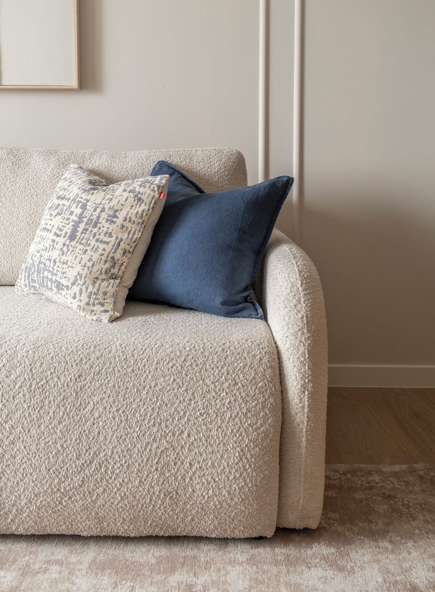 A beige sofa with two pillows, one patterned and one solid blue, against a plain wall and wooden floor.