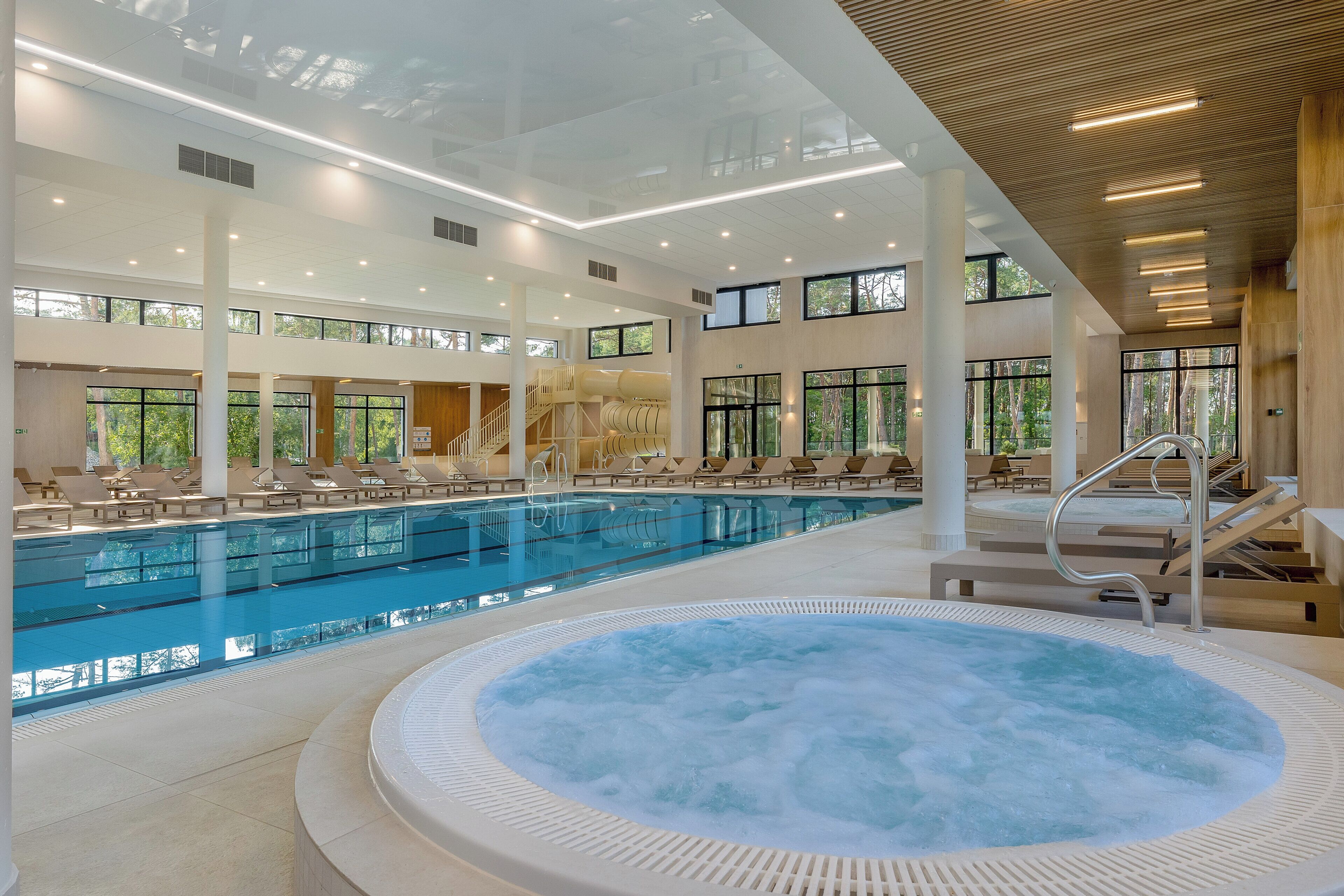 A large indoor swimming pool with a hot tub, surrounded by lounge chairs and a staircase leading to upper levels.