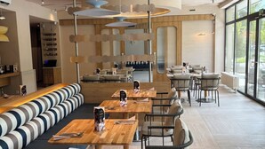 Un interior de restaurante moderno con mesas de madera, asientos acolchados, lámparas colgantes y ventanas amplias.