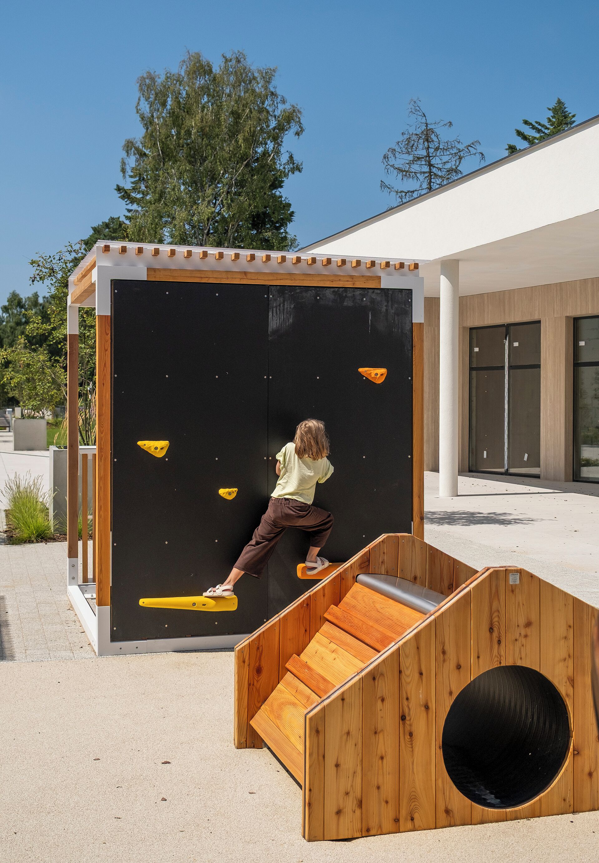 Ein Kind erklimmt eine Holz-Kletterwand mit schwarzem Hintergrund und gelben Griffen.
