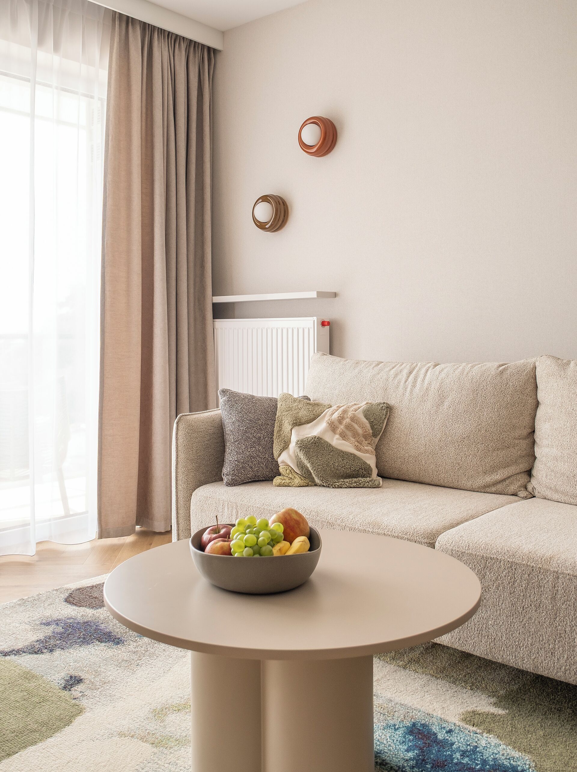 A living room with a beige sofa, a round table with a bowl of fruit, and a radiator on the wall.