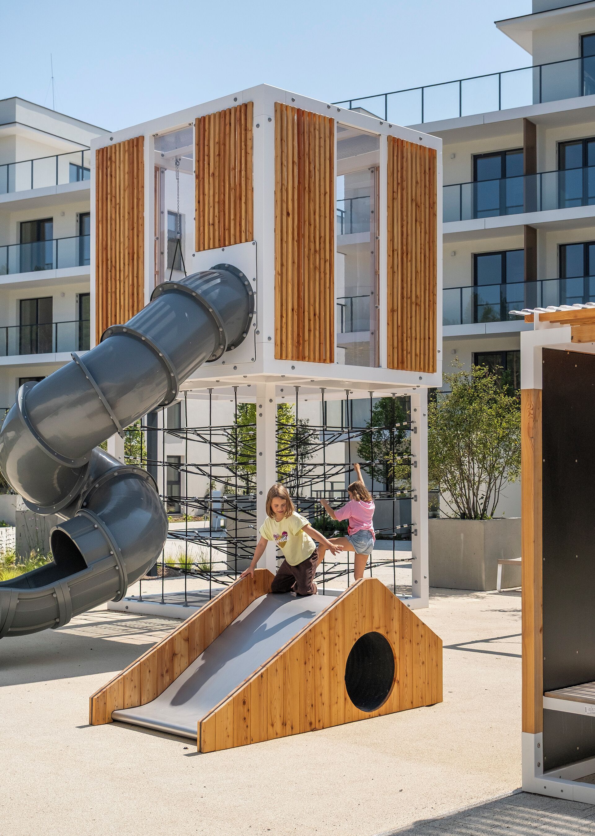 A playground with a slide and climbing structure, surrounded by modern buildings.