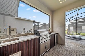 Eine Außenküche mit Edelstahlgrill, Spüle und Fenster mit Blick auf einen Garten.
