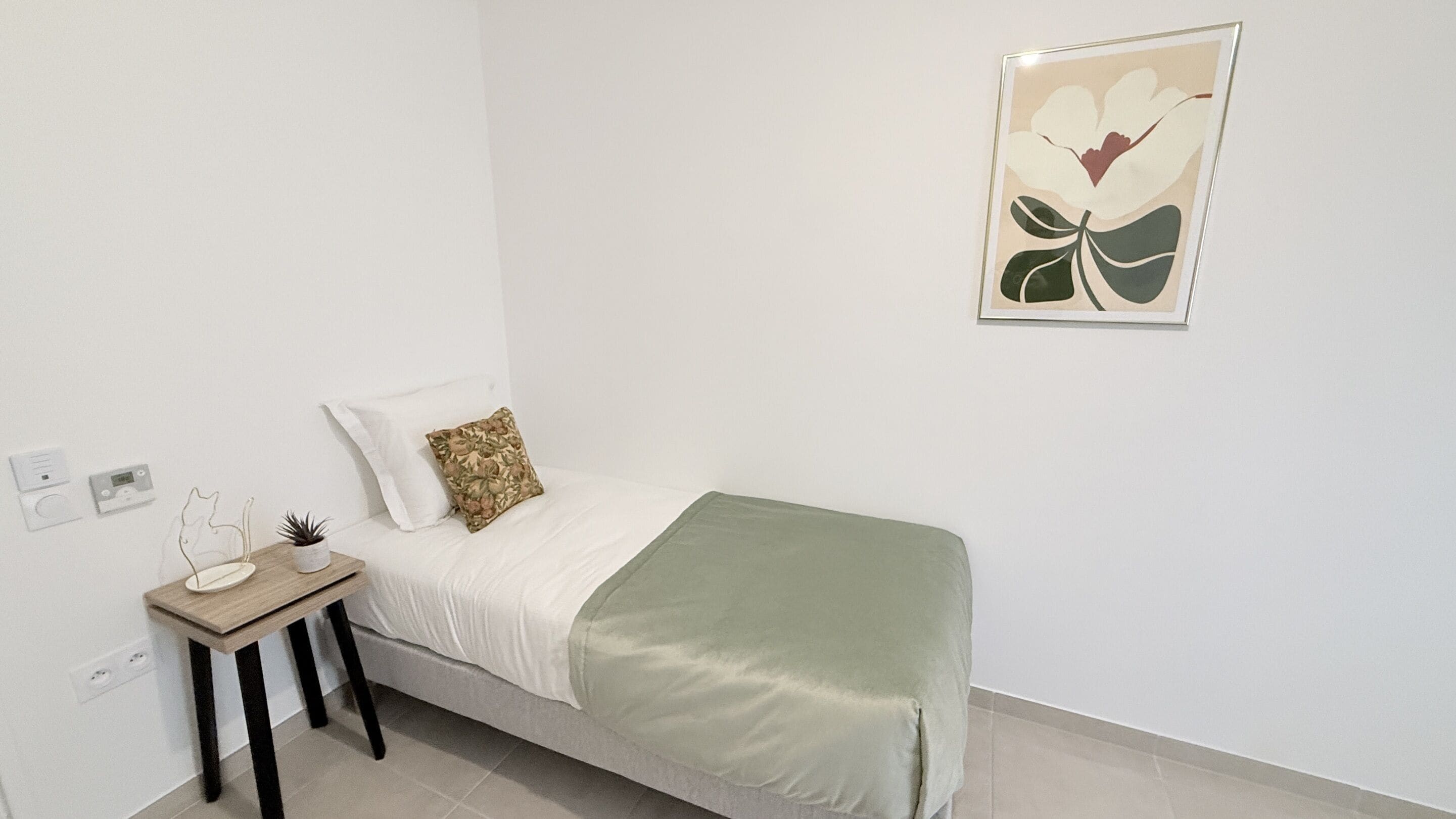 A bedroom with a bed, a bedside table, a small plant, a pillow, a blanket, and a framed picture on the wall.