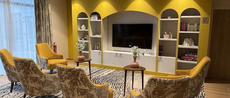 A living room with yellow walls, a large flat-screen TV, and multiple bookshelves. There are three armchairs and a small table with a vase.