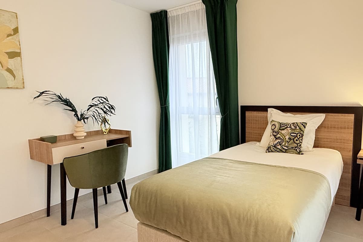 Une chambre d’hôtel comprenant un lit, un bureau avec une chaise, une table de chevet avec une lampe et une fenêtre avec des rideaux.