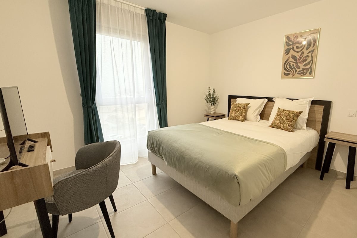Une chambre à coucher comprenant un lit, une chaise, un bureau, une table de chevet et une fenêtre avec des rideaux.