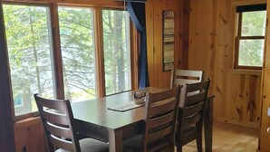 A wooden cabin interior with a dining table and chairs, large windows, and a sign indicating a cabin rental. - Cozy 2 bedroom cottage on lake! (Mountain)