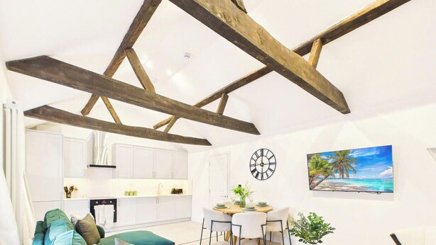 A modern kitchen with white cabinets, a dining area with a wooden table and chairs, and a large clock on the wall. - *NEW* Artisan Apartment | Modern | Spacious (Gloucestershire)