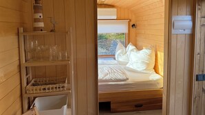 Innenansicht einer Holzhütte mit einem Bett, Regalen mit Gläsern, einem Korb und einem kleinen Zierturm.