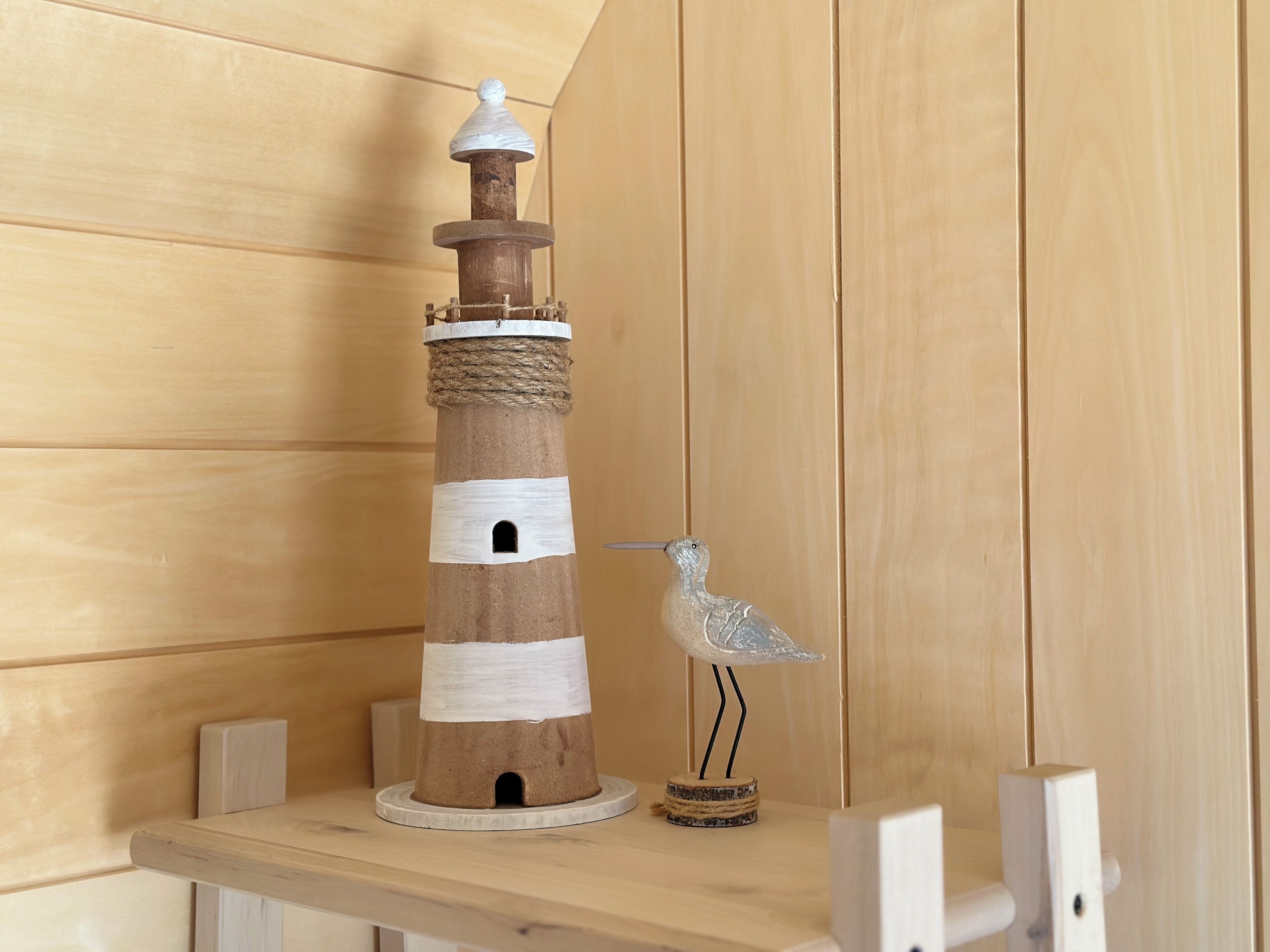 Un phare en bois avec une corde enroulée autour de sa base, une figurine d’oiseau posée sur un petit socle en bois, et une étagère sur laquelle se trouve un objet blanc.
