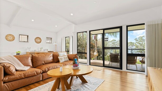 A spacious living room with a brown leather sofa, a wooden coffee table, and a large sliding glass door leading to an outdoor area with wicker furniture. - Roya by the Beach by Experience Jervis Bay (Vincentia)