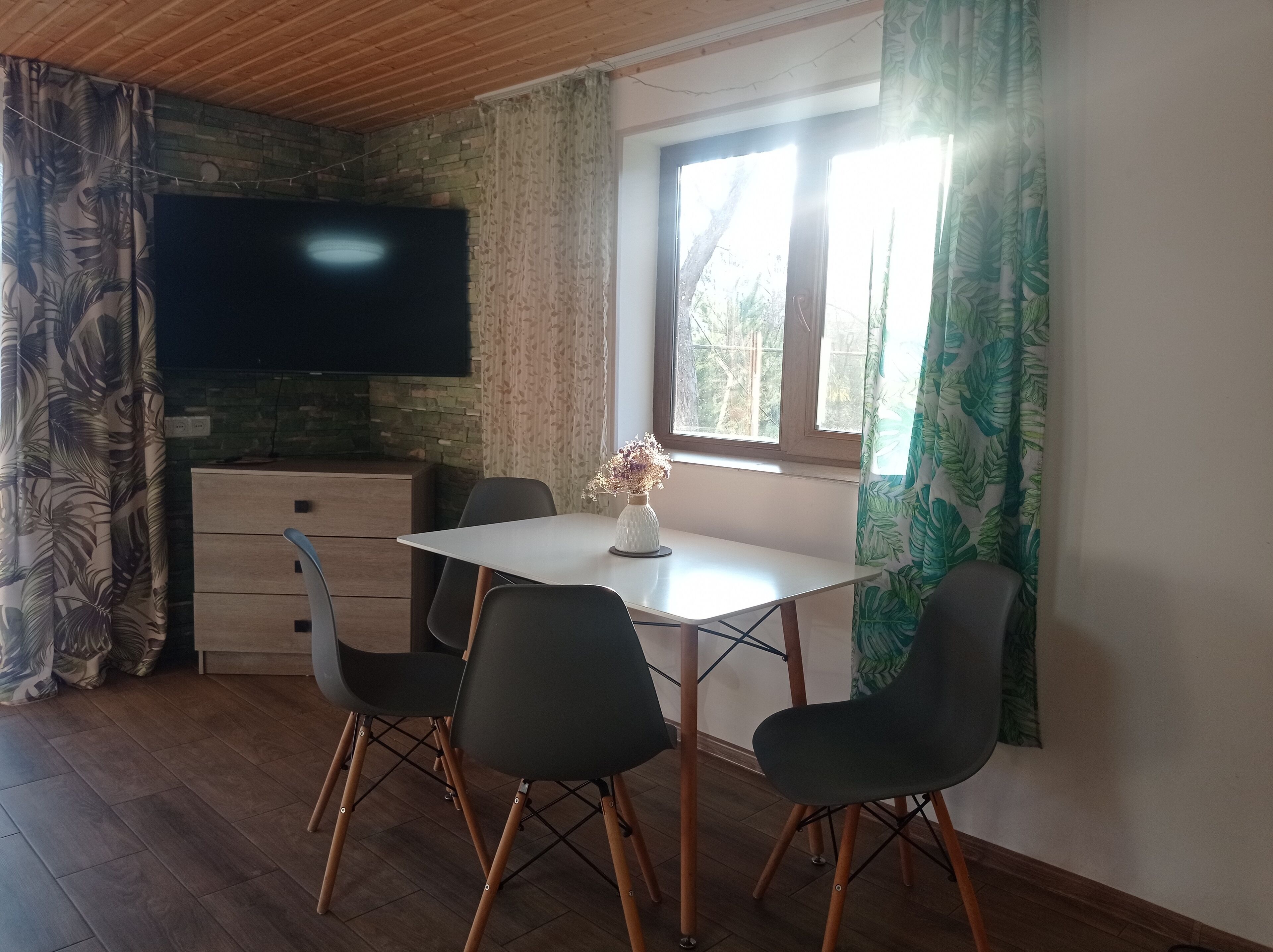 Un coin repas avec une table et des chaises, un téléviseur à écran plat fixé au mur et une fenêtre avec des rideaux.