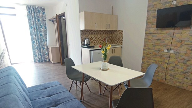 A modern living room with a blue sofa, a dining table with chairs, and a kitchen area with a stone backsplash and a flat-screen TV mounted on the wall. - Amrakits Canyon Guesthouse (Amrakits)