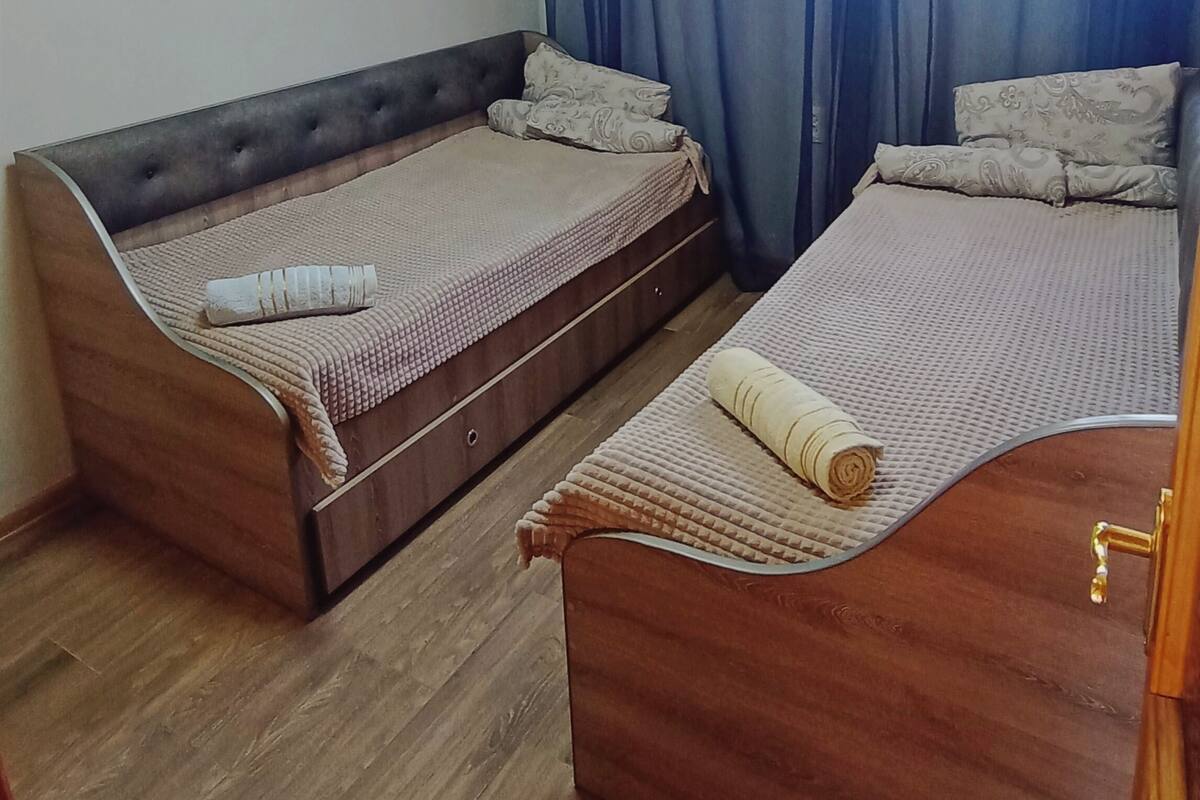 A room with two wooden beds, a window with blue curtains, and a wooden ceiling.