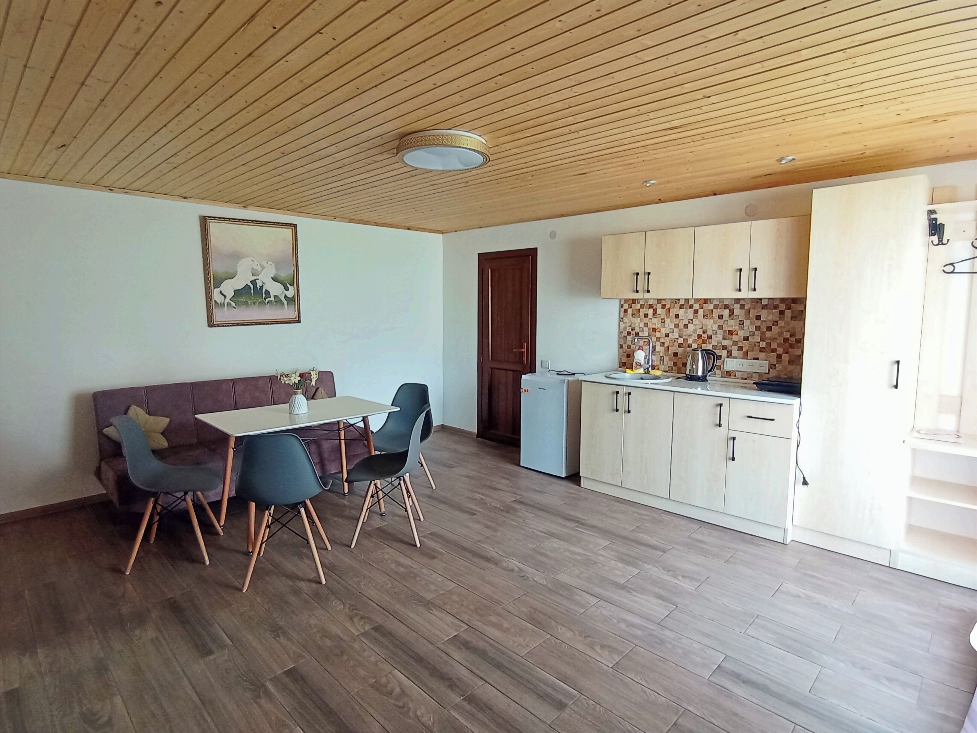 A dining area with a wooden ceiling, a painting on the wall, a dining table with chairs, a sofa, and a kitchen area with cabinets and a countertop.