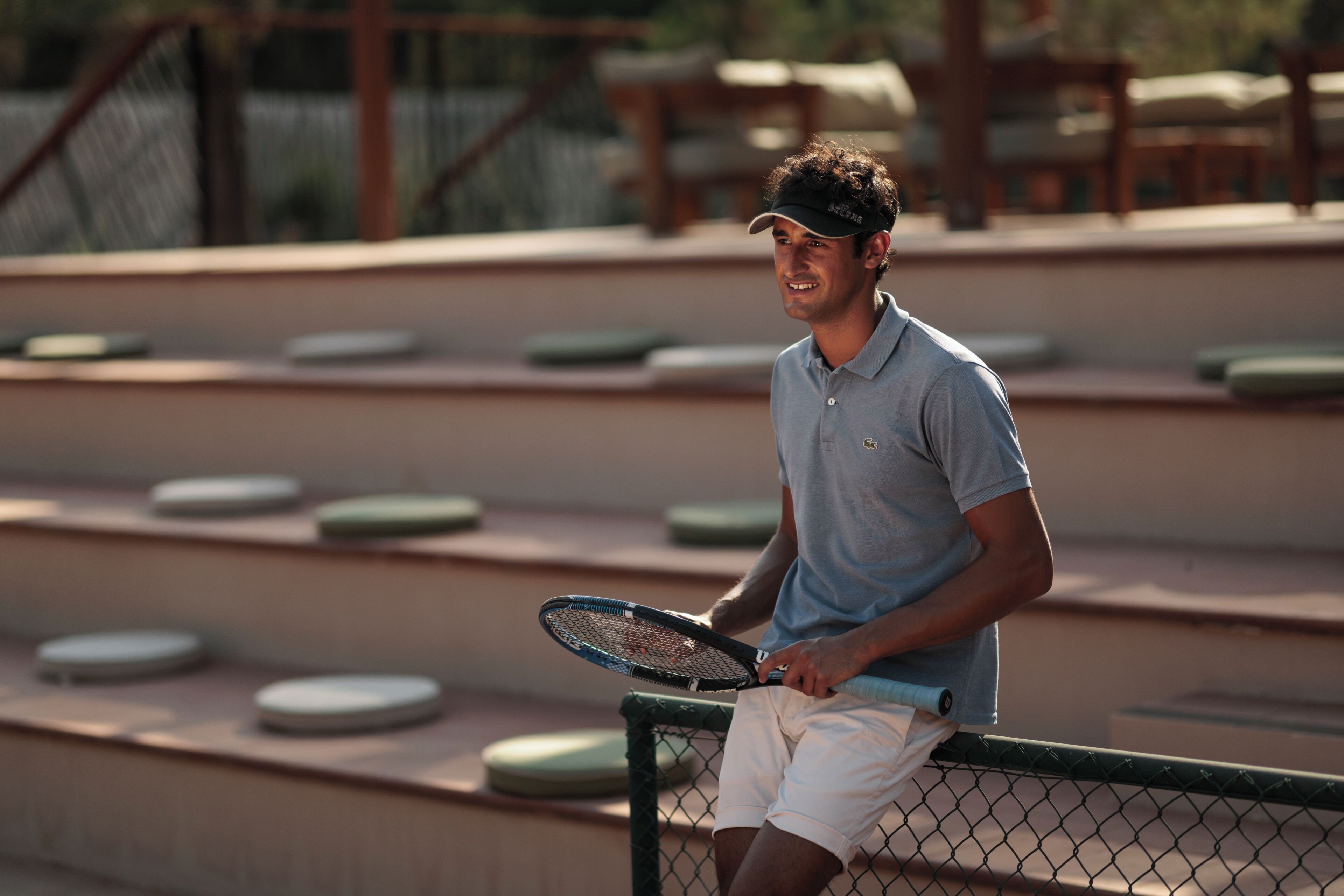 A tennis player holding a racket, seated on a tennis court with a chain-link fence and tiered seating in the background.