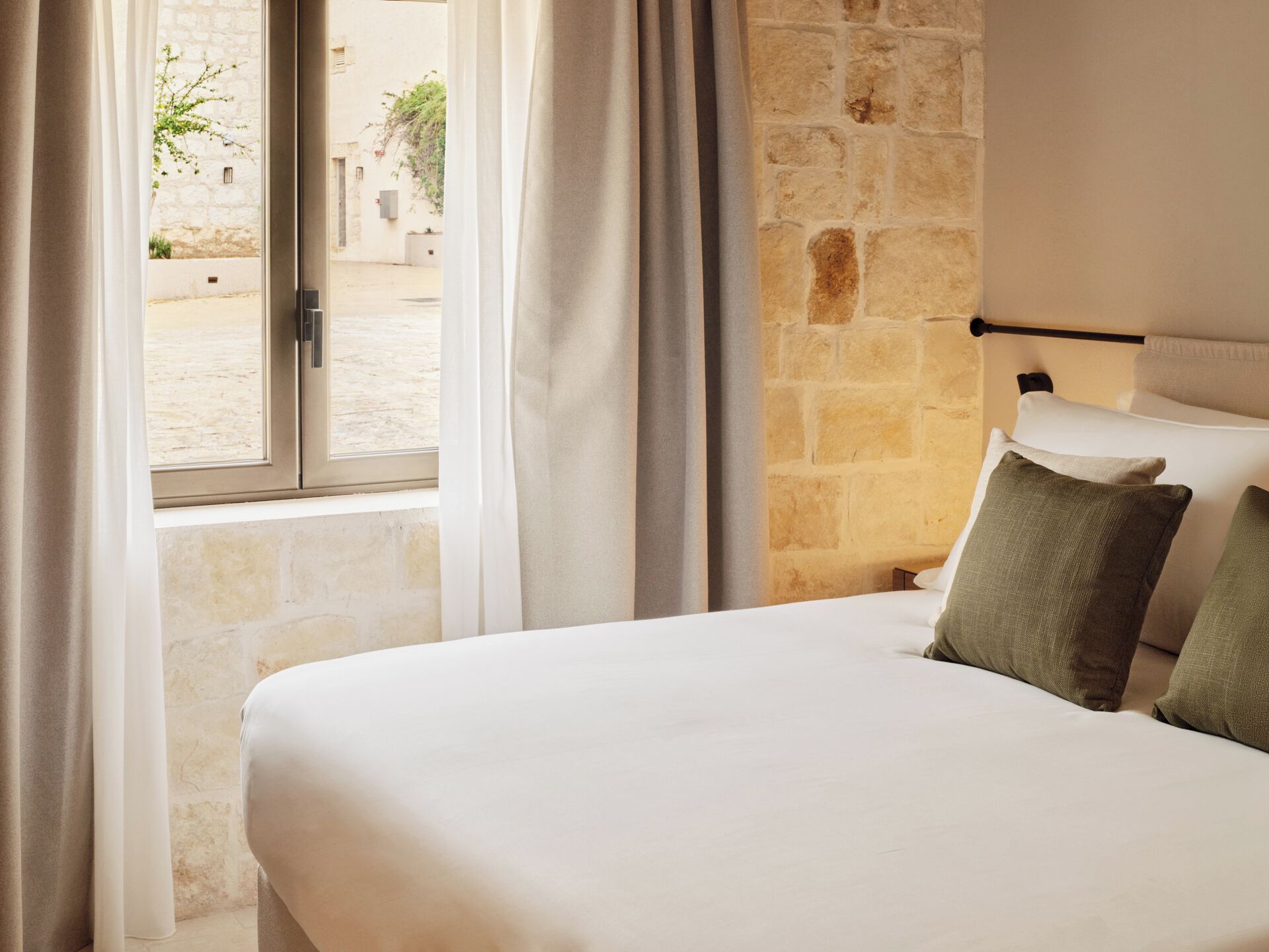 A hotel room with a bed, curtains, a stone wall, and a window with a view of an outdoor area.