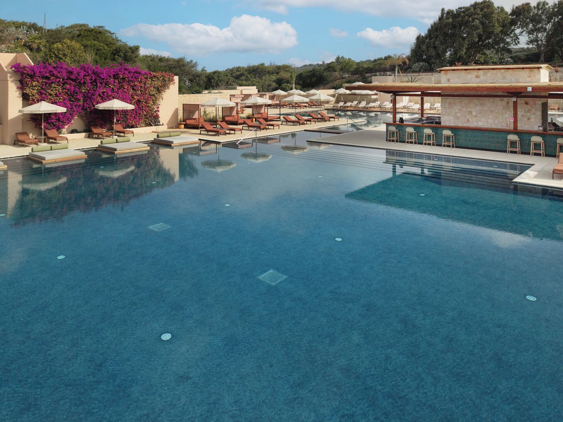 A large, clear swimming pool with lounge chairs and umbrellas, surrounded by greenery and a bar area.