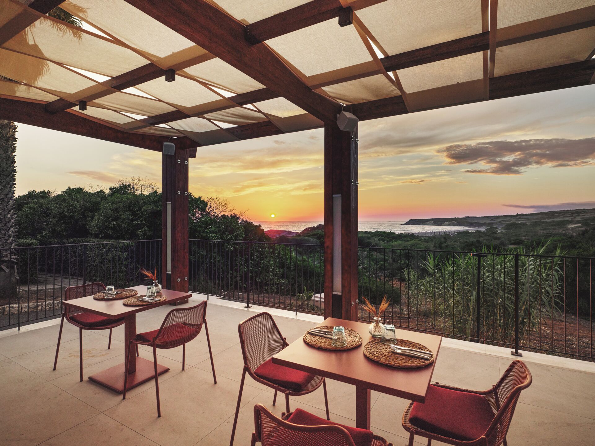 An outdoor patio with tables and chairs, a view of the sunset, and a railing.