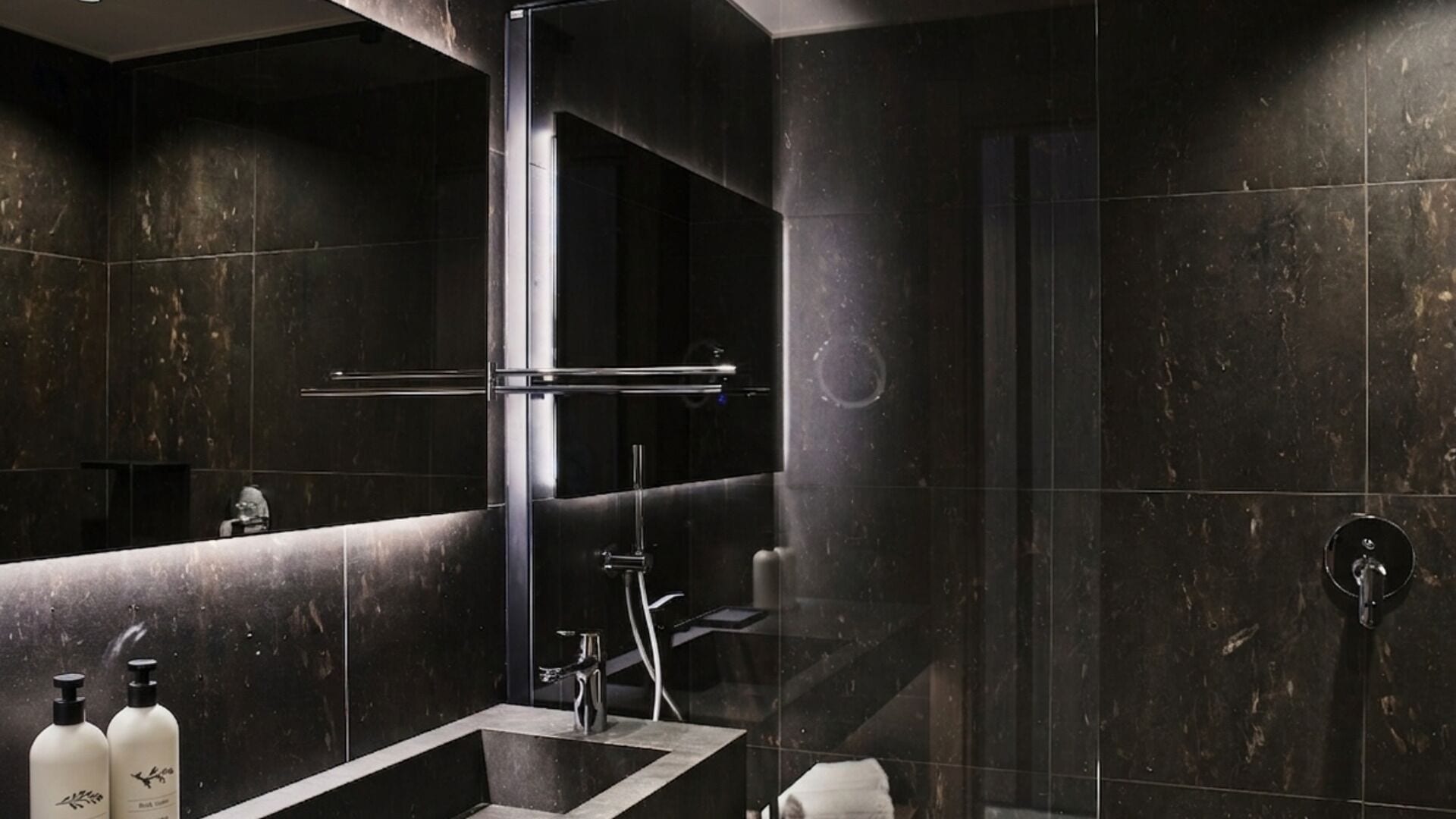 A modern bathroom with dark marble tiles, a glass shower enclosure, a vanity with a mirror, and two bottles of shampoo.