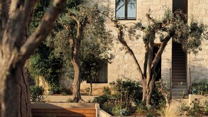 Un bâtiment en pierre, doté d’un toit en tuiles, entouré d’arbres adultes et de parterres aménagés.