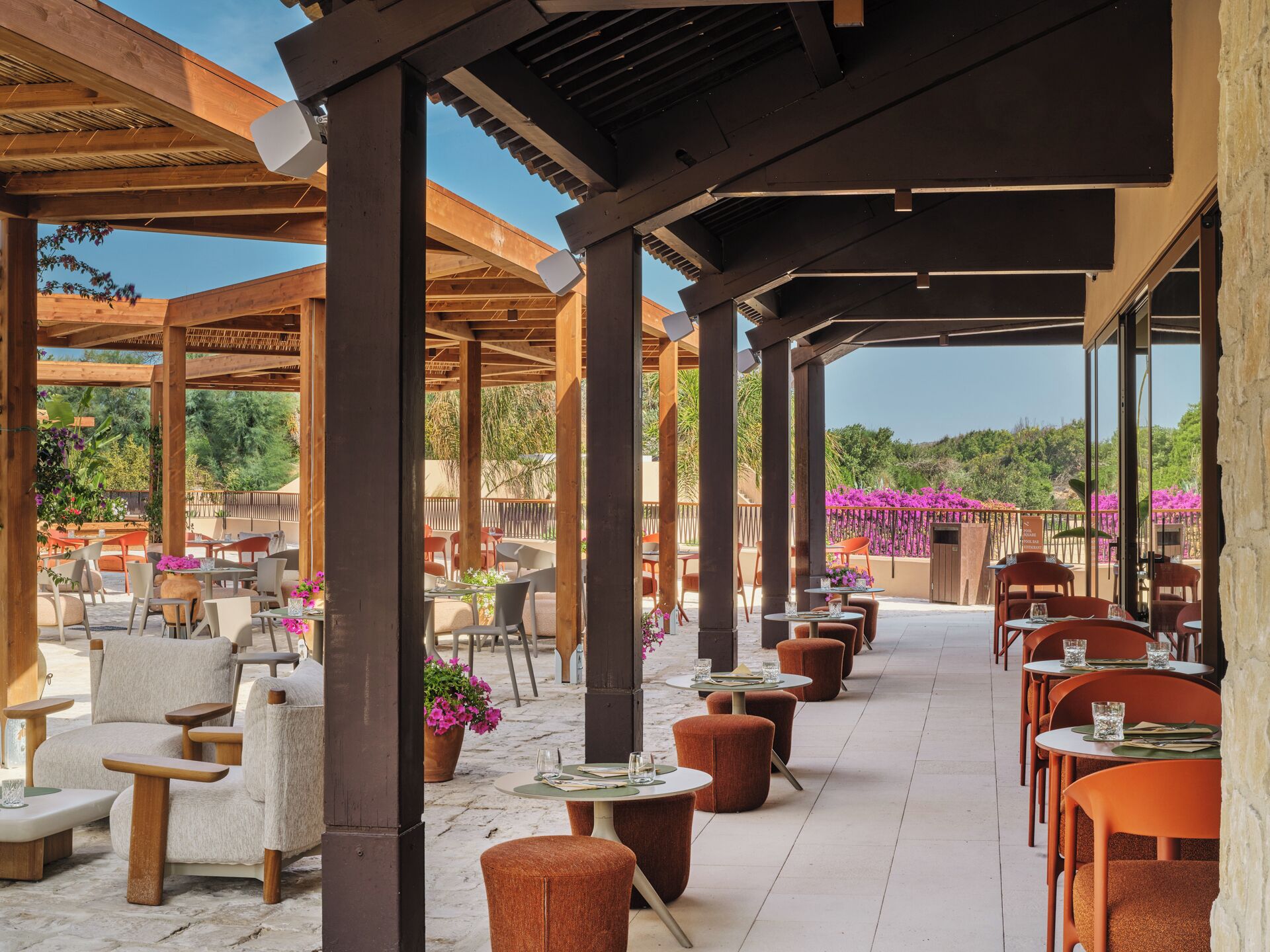 Una terrazza esterna con tavoli e sedie di legno, immersa nel verde e tra fiori.