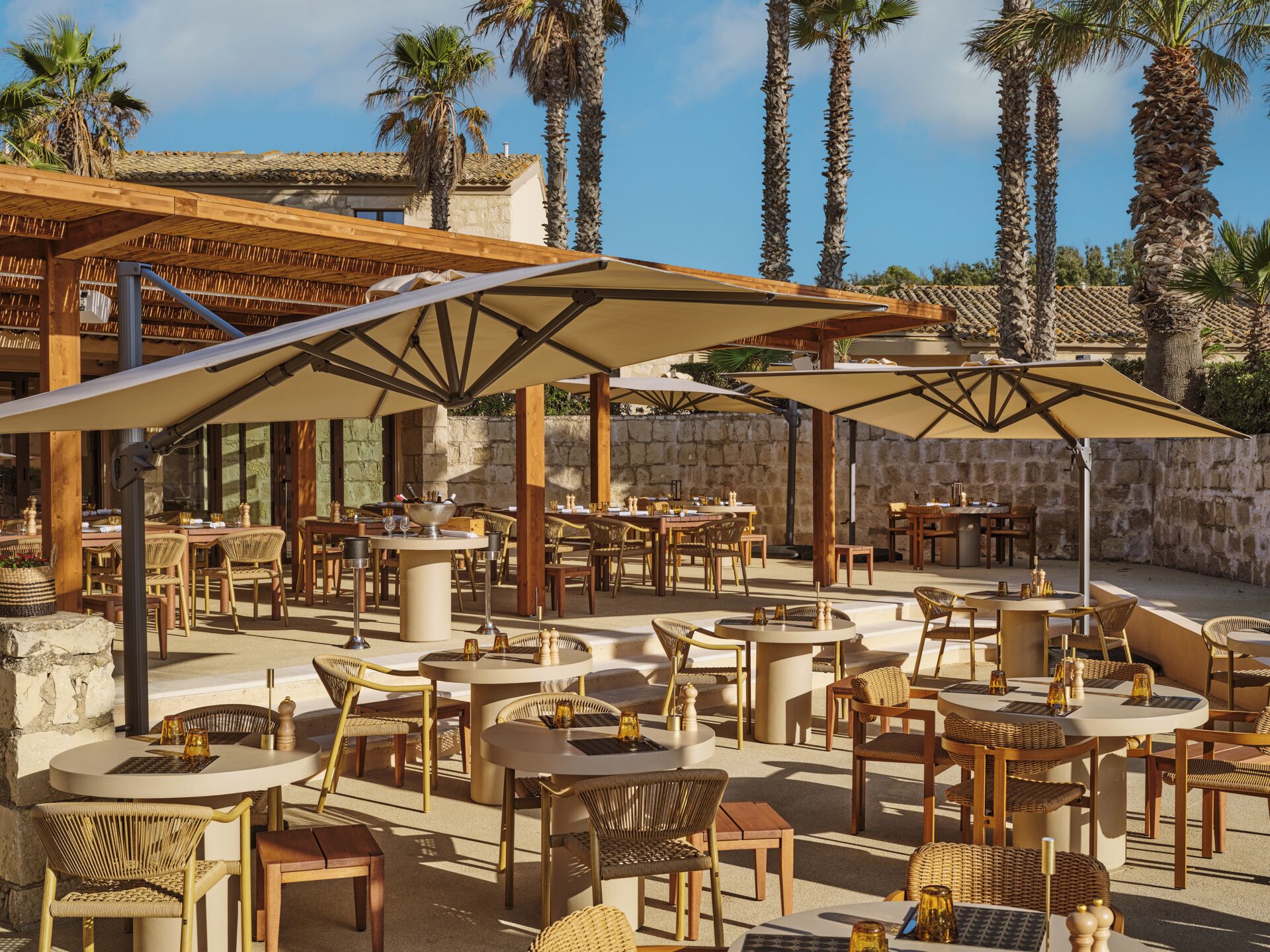An outdoor patio with round tables and chairs, umbrellas, and palm trees.