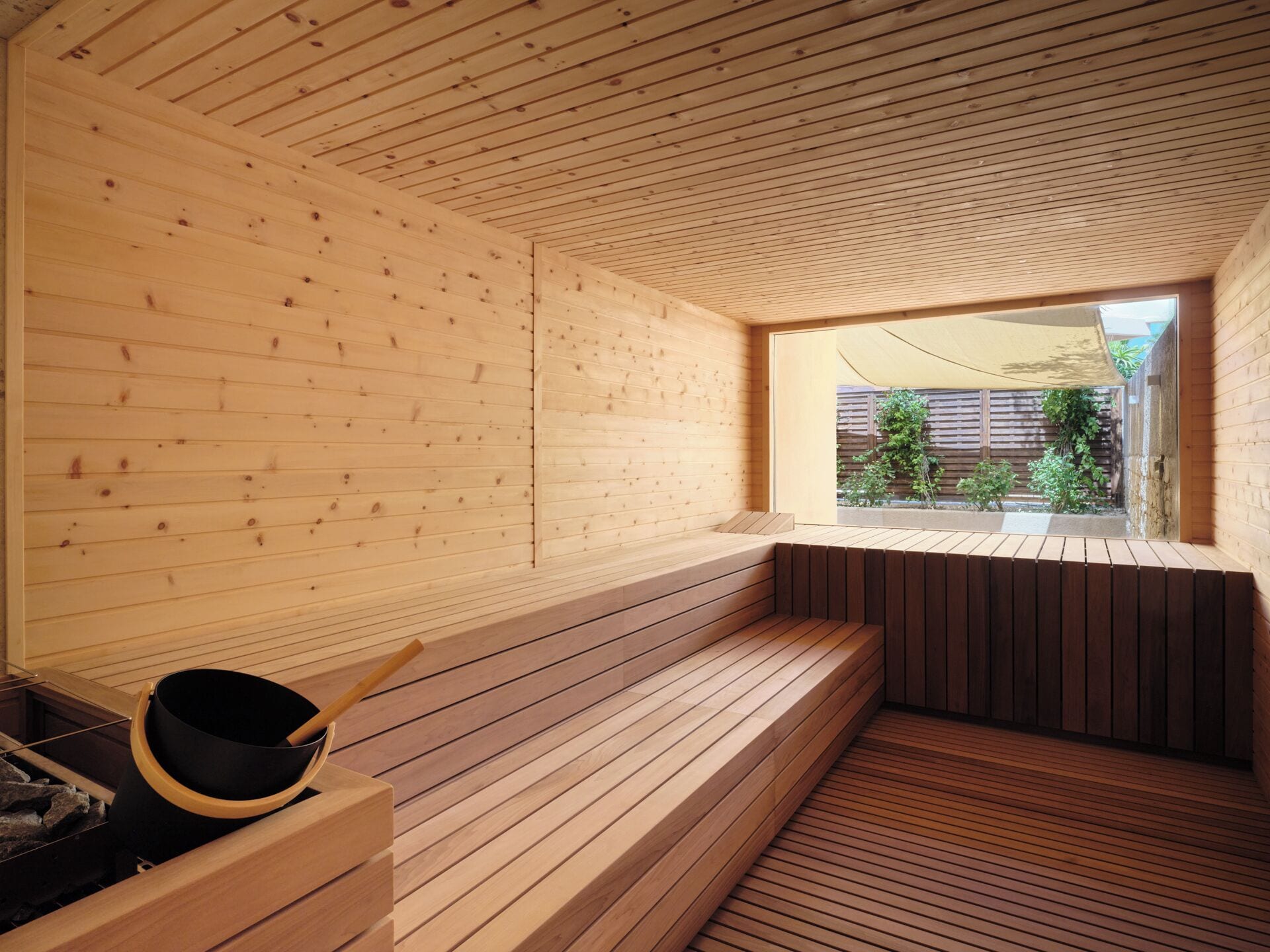 A sauna room with wooden walls and benches, a window with a view of greenery, and a black container with a wooden handle.