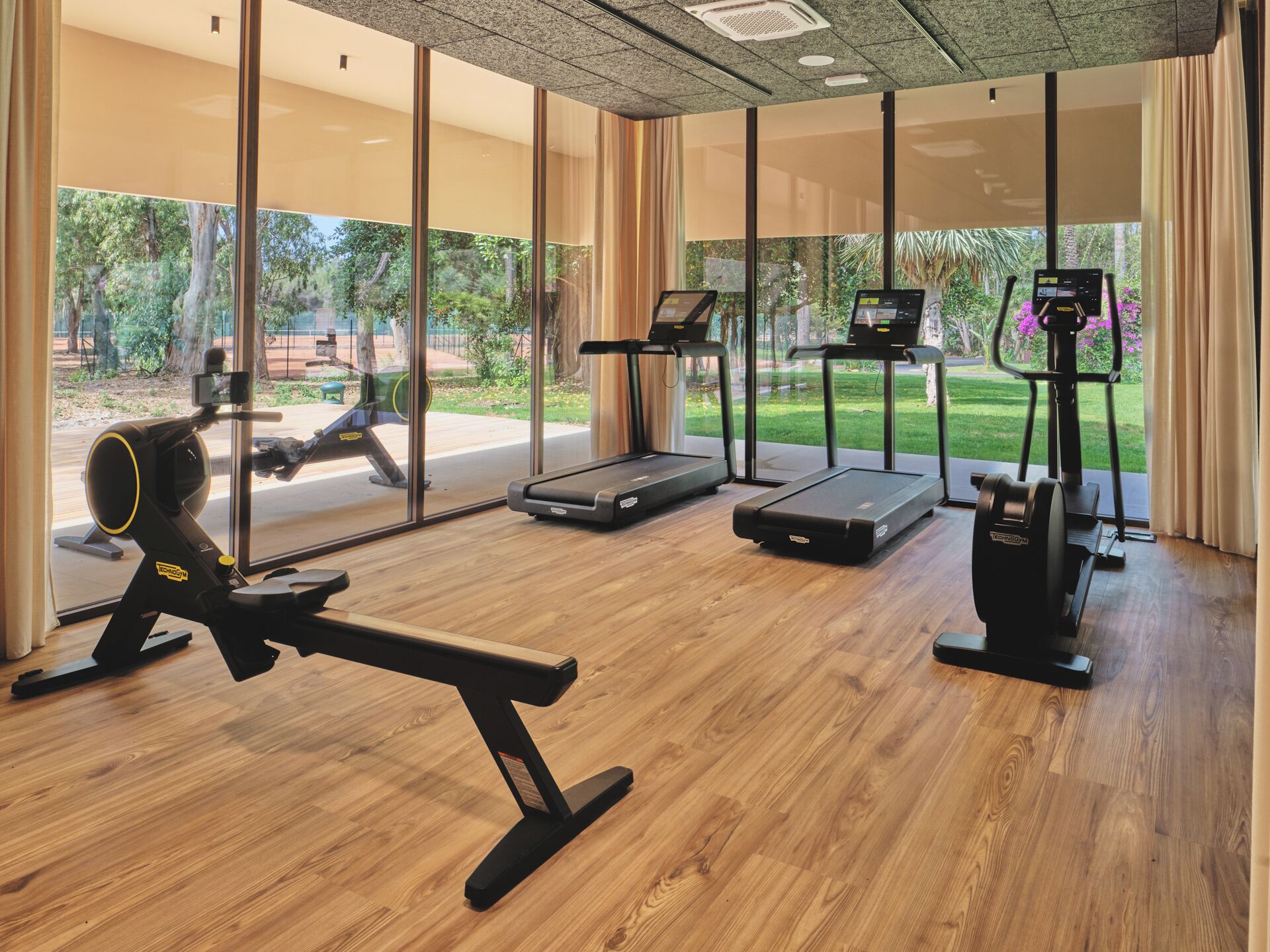 A gym with treadmills, a rowing machine, and a stationary bike, with large windows offering a view of the outdoors.