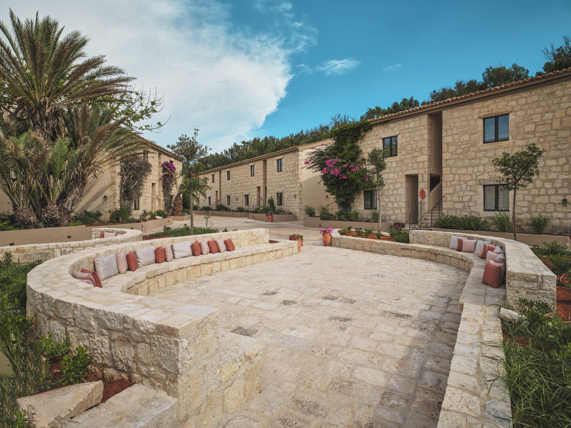 A courtyard with stone benches, a palm tree, and stone buildings with windows.