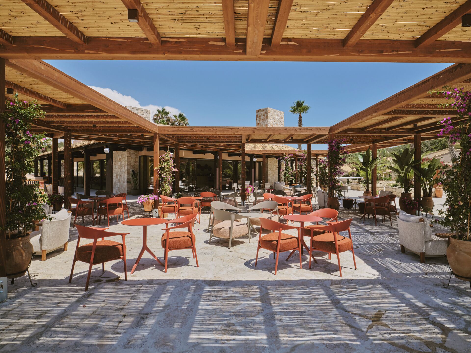 Un patio exterior con mesas y sillas de madera, rodeado de plantas en macetas y un muro de piedra.