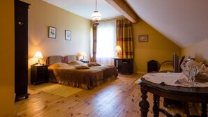 A cozy bedroom with a wooden floor, a bed with a headboard, a nightstand, a lamp, a window with curtains, and a dining table with a glass. - SPA Hotel Mārcienas Muiža (Mārciena)