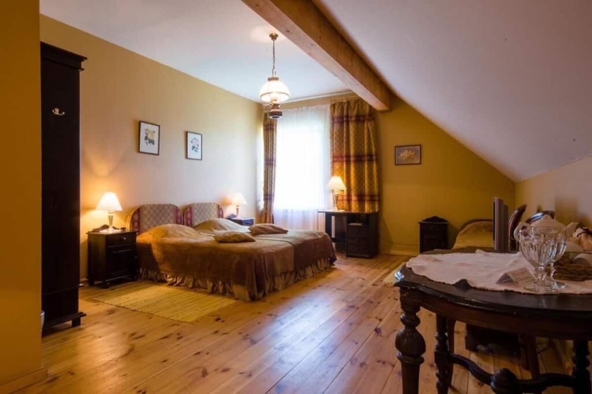 A cozy bedroom with a wooden floor, a bed with a headboard, a nightstand, a lamp, a window with curtains, and a dining table with a glass.