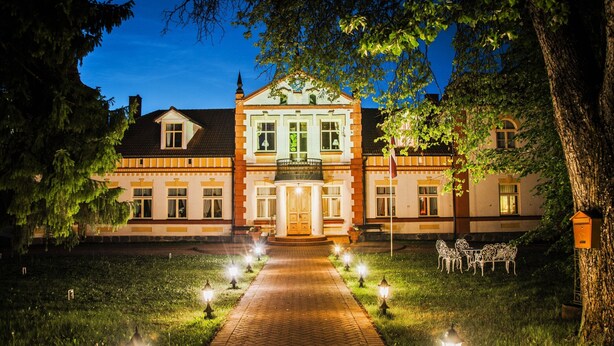 A large, historic building with a central entrance, surrounded by a well-lit garden and outdoor seating area. - SPA Hotel Mārcienas Muiža (Mārciena)