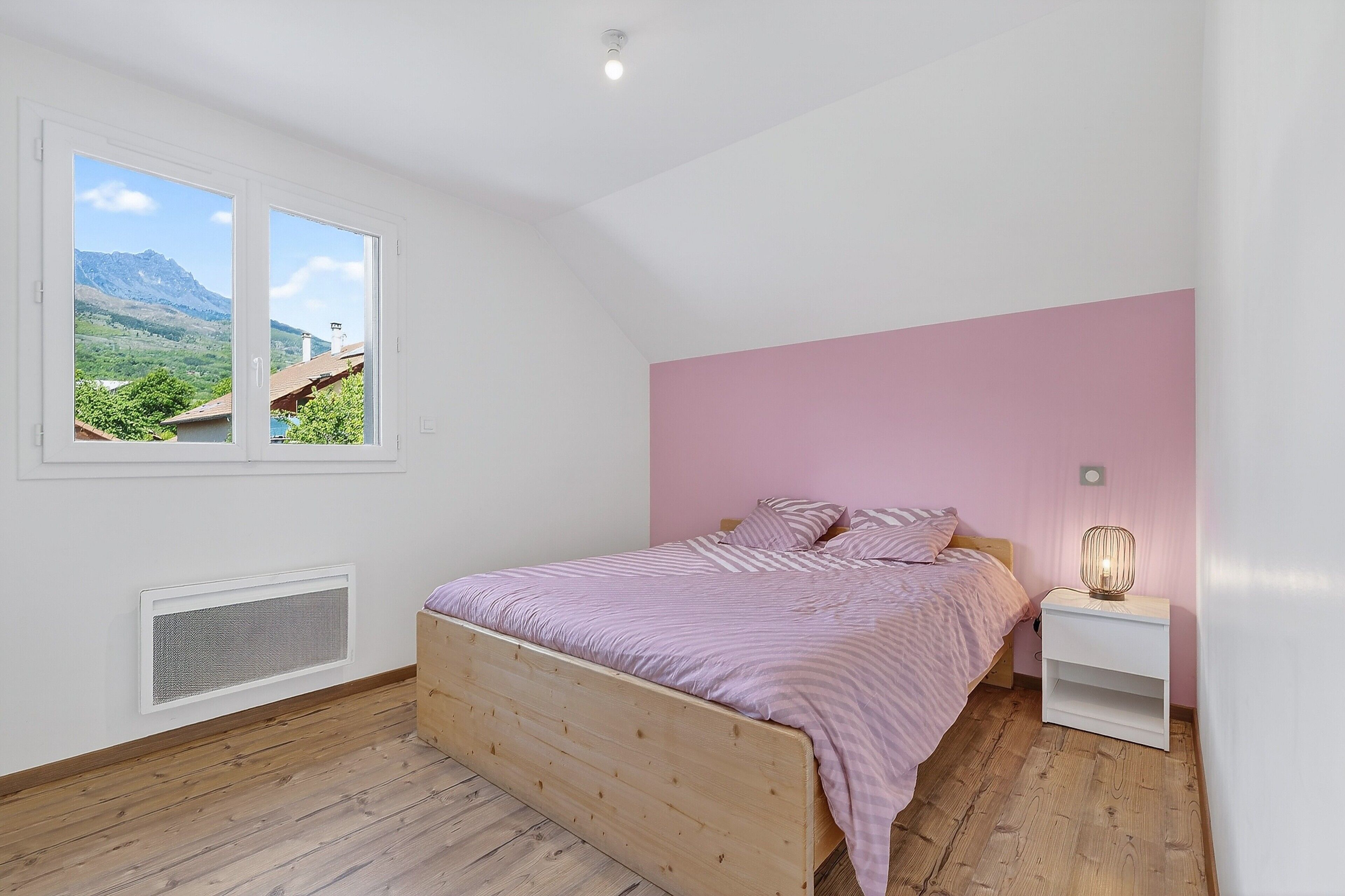 A bedroom with a wooden bed, a nightstand, a window with a view of mountains, and a pink wall.