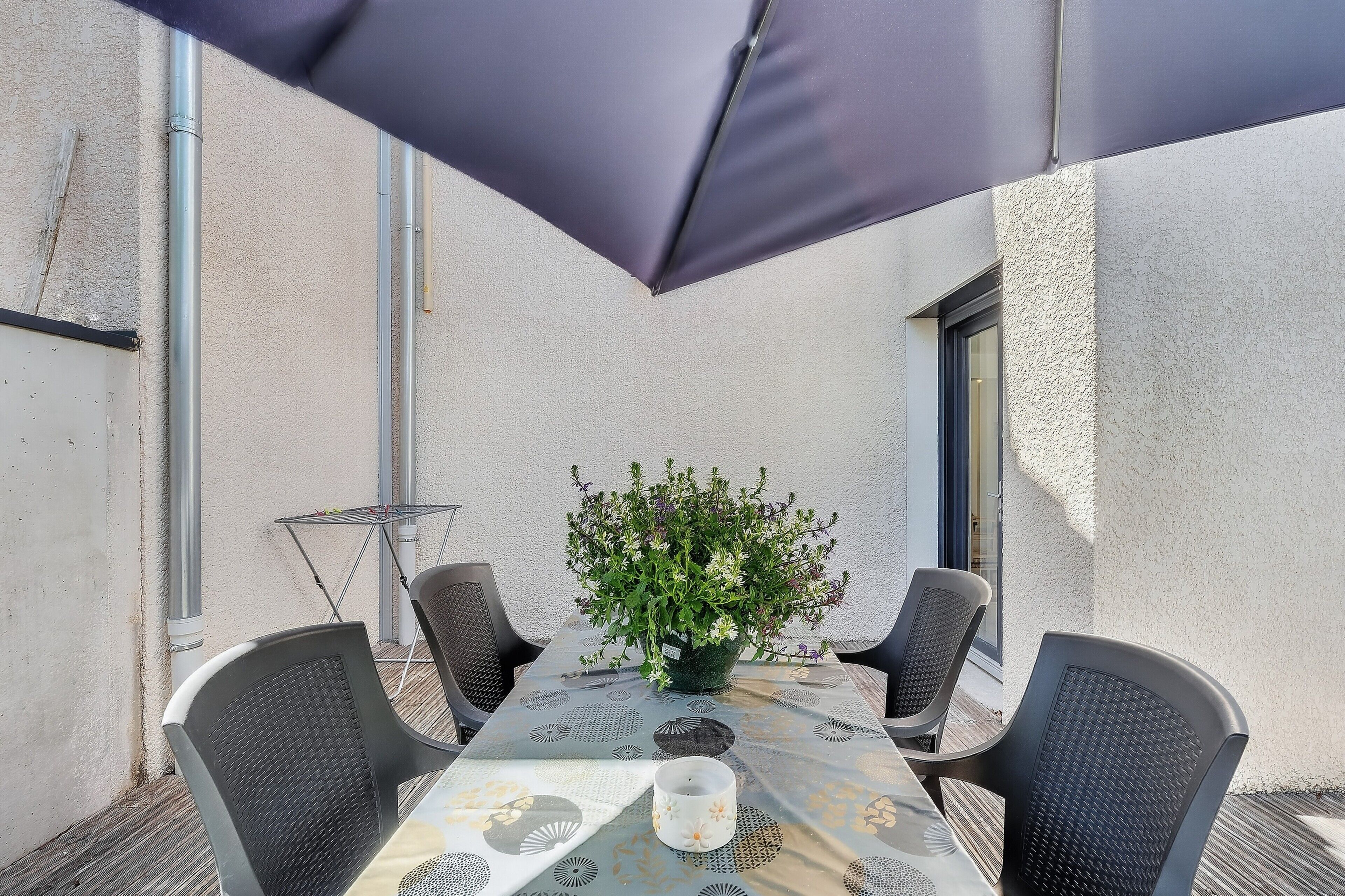 Une terrasse extérieure avec une table, des chaises, un pot de fleurs et un grand parasol.