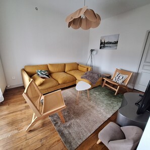 Ett vardagsrum med en soffa i senapsgul färg, ett litet vitt sidobord, en grå matta, en trästol och en golvlampa.