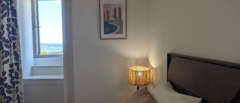 A bedroom with a bed, pillows, a bedside lamp, a window with a view, and a framed picture on the wall.